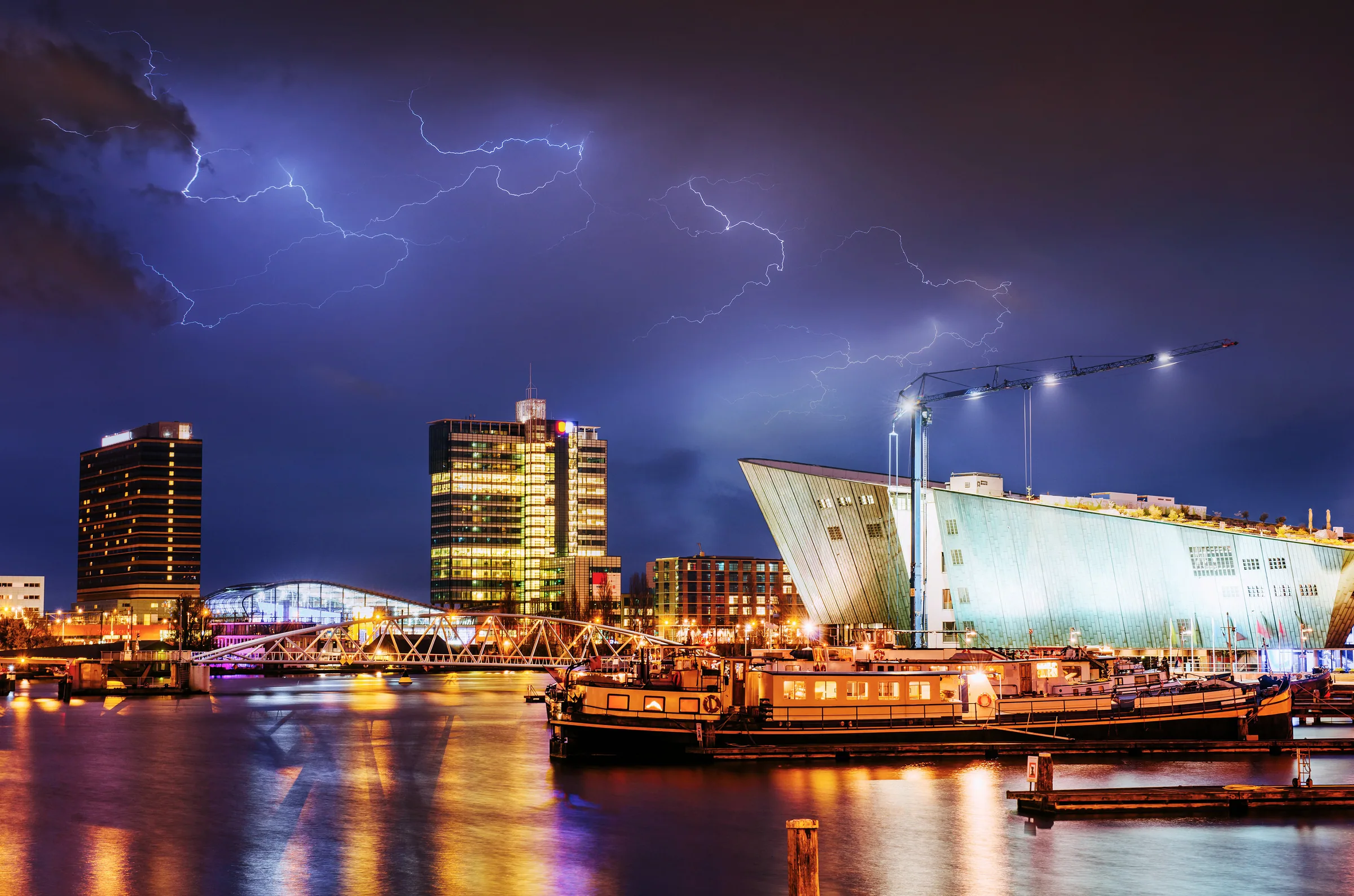 Amsterdam nightlife guide — the Oosterdok at night under a lightning sky, the NEMO roof and the harbour towers lit above the water on the eastern docks where Panama, the city's most event-listed club, sits