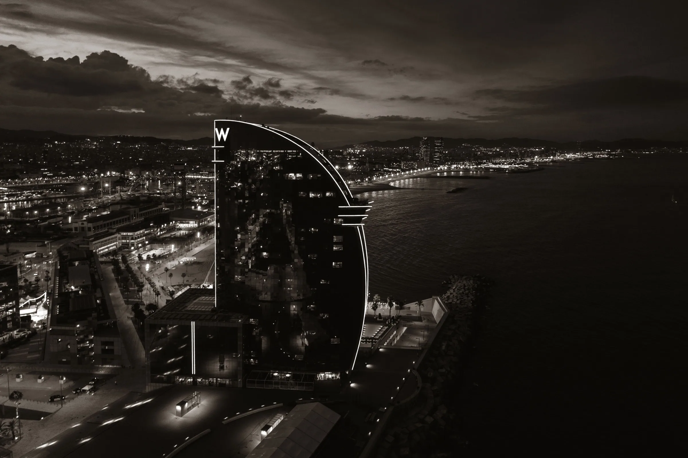 Barcelona nightlife guide — the W Hotel on the Barceloneta seafront from the air at dusk, the Passeig Marítim beachfront lights running north toward Opium and Shôko, the two most expensive club doors in the city