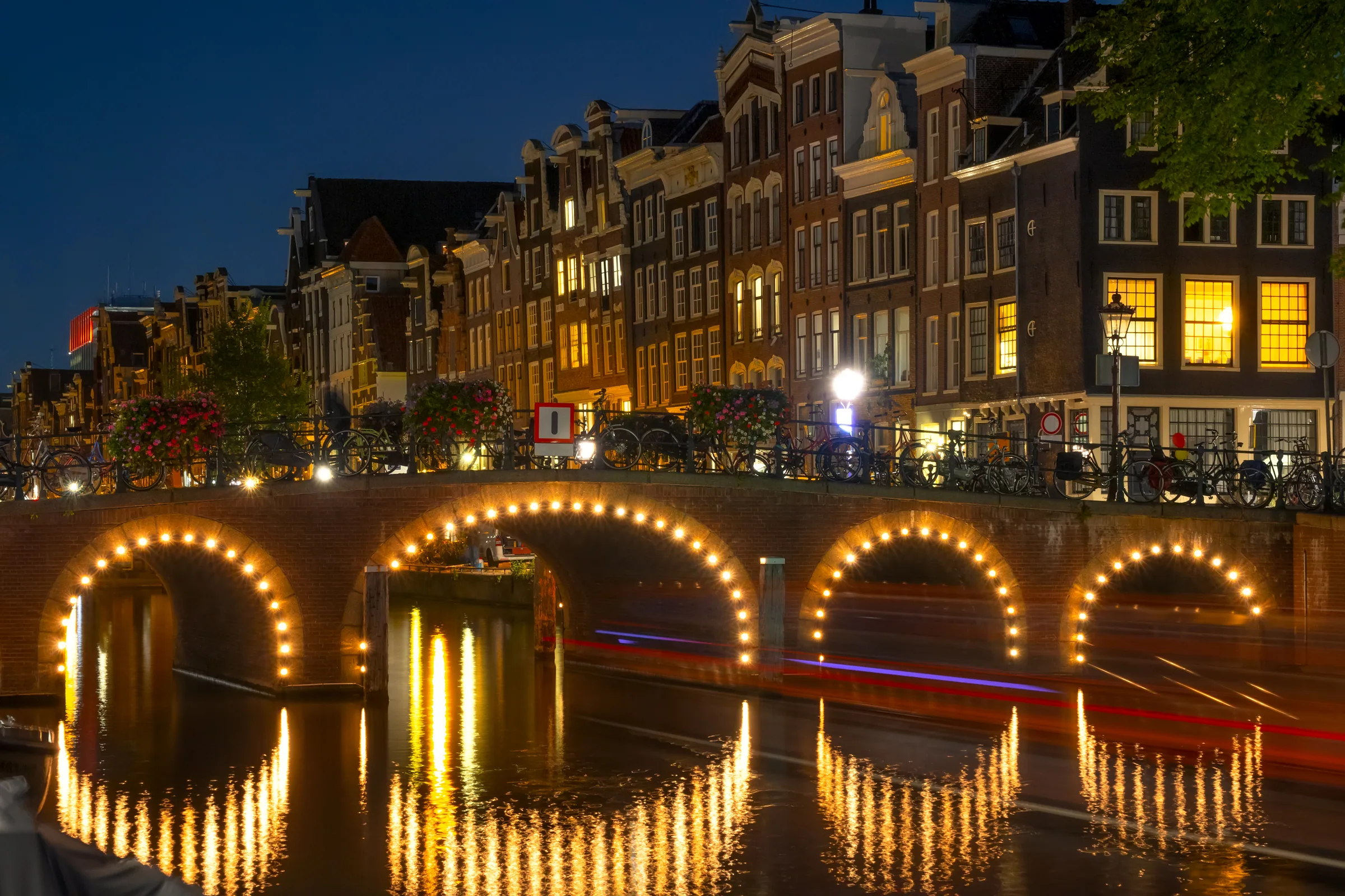 Best nights to go out in Amsterdam — the lit arches of a canal-belt bridge at blue hour, boat light trails streaking under the spans, the Grachtengordel between Leidseplein and Rembrandtplein on a night in progress