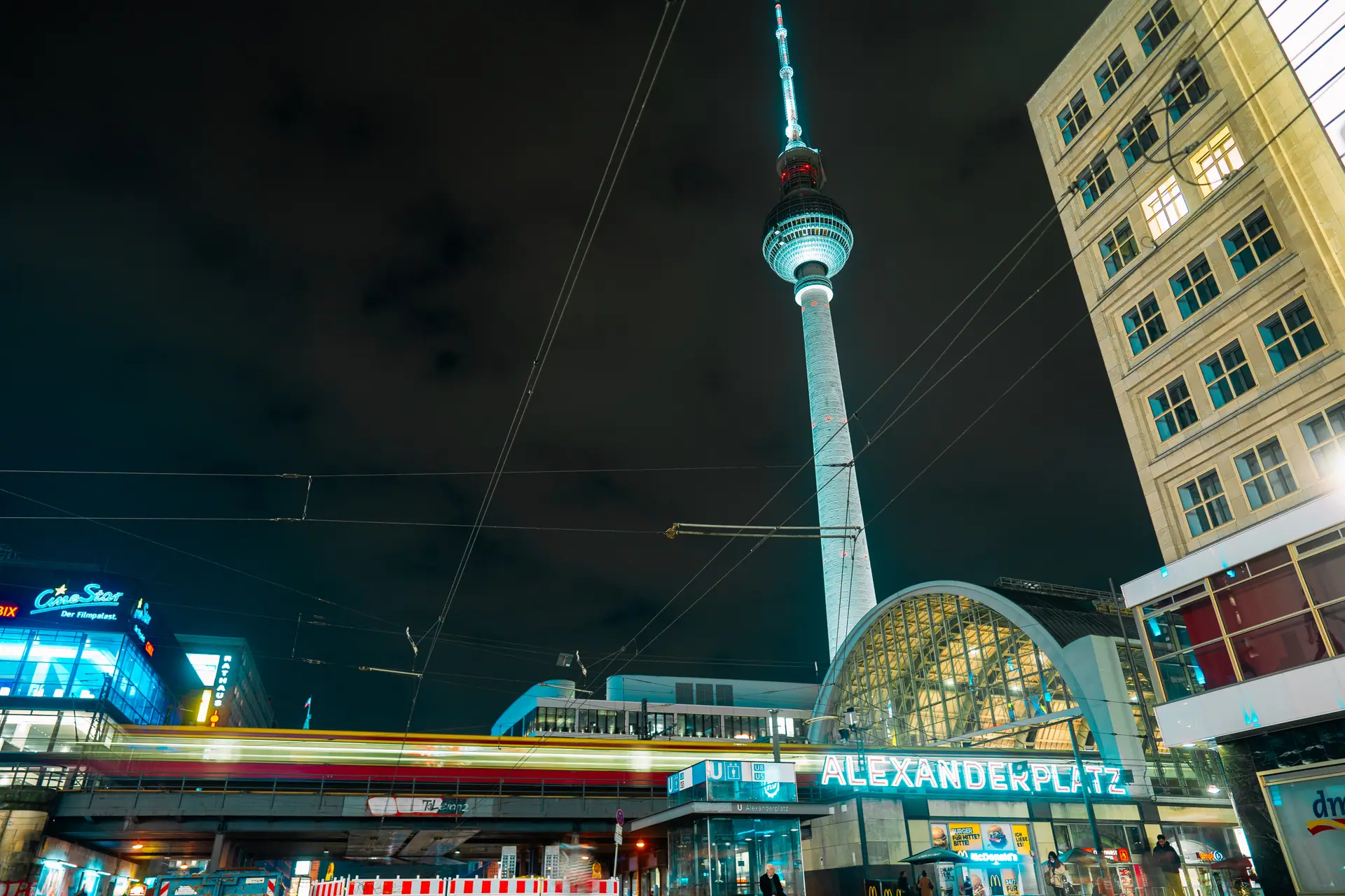 Clubs in Berlin — Alexanderplatz at night with the Fernsehturm lit above the station and a tram streaking past, a block from the clubs at Alexanderstraße 7