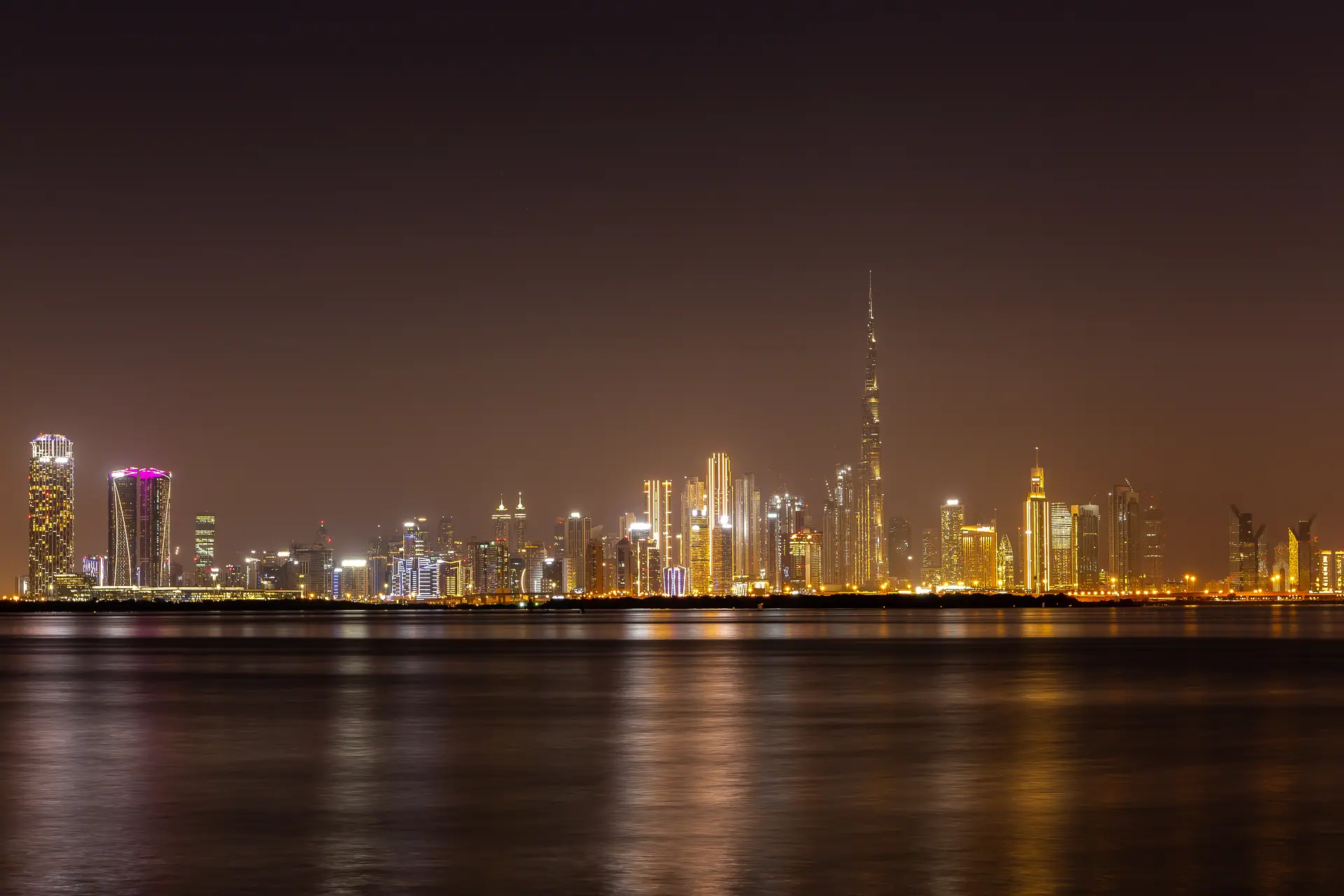 Dubai nightlife — the Downtown and Business Bay skyline lit at night across the water, the Burj Khalifa in the center, the Sheikh Zayed Road hotel towers that hold most of the city's licensed bars and clubs