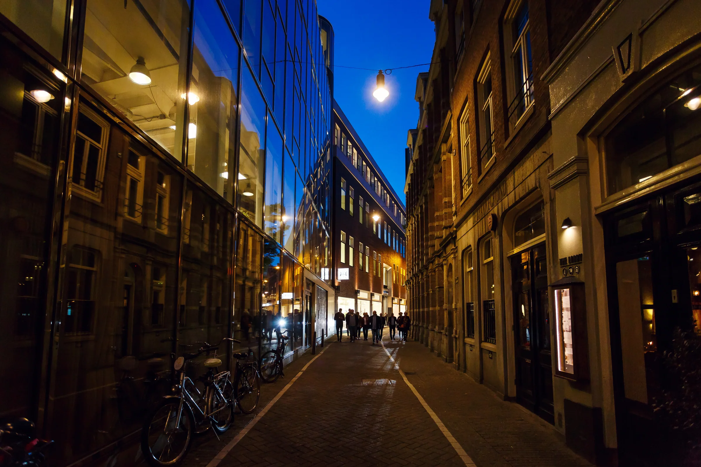 Parties in Amsterdam — a group walking down a central Amsterdam side street at blue hour, bikes against the glass and the lit shopfronts at the end of the lane